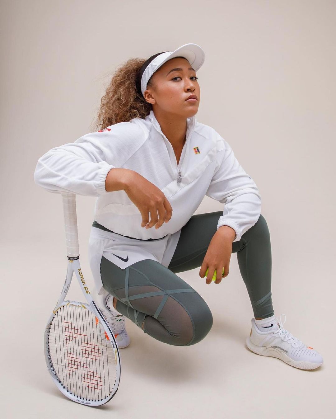 Athlete Naomi Osaka in athletic wear poses with a tennis racket and tennis ball in a studio environment against a neutral backdrop.