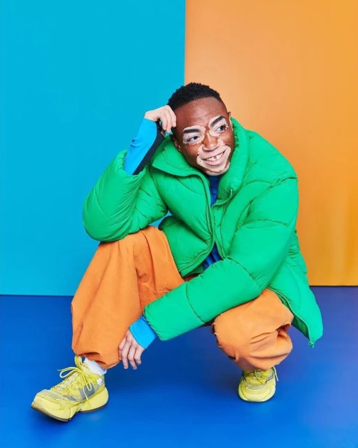 Portrait of a man with vitiligo wearing a green puffer jacket and orange pants, crouching against a backdrop of blue and orange.