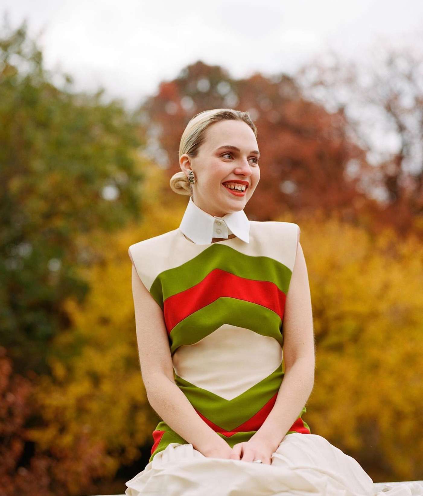 Portrait of a blonde woman in a cream dress with green and red chevron stripes sitting in front of trees with autumn foliage.