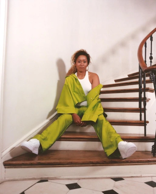 A woman in a bright green outfit sits on wooden stairs in a relaxed pose, with a white wall and black and white tile floor.