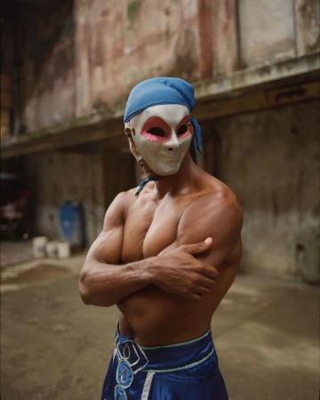 Shirtless man in a mask and blue head wrap stands with arms crossed against a weathered building in an urban setting.