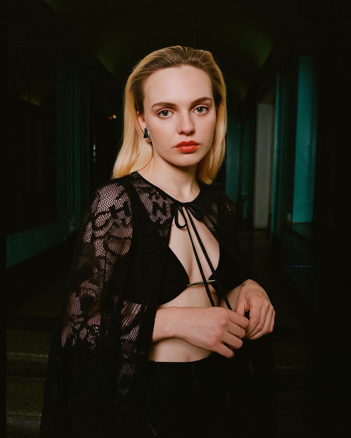 Portrait of a blonde woman in a lace top and black pants standing in a dimly lit hallway looking directly at the camera.