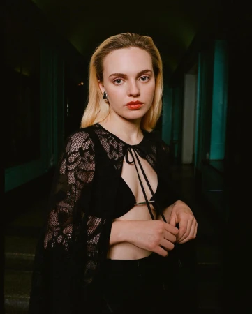 Portrait of a blonde woman in a lace top and black pants standing in a dimly lit hallway looking directly at the camera.