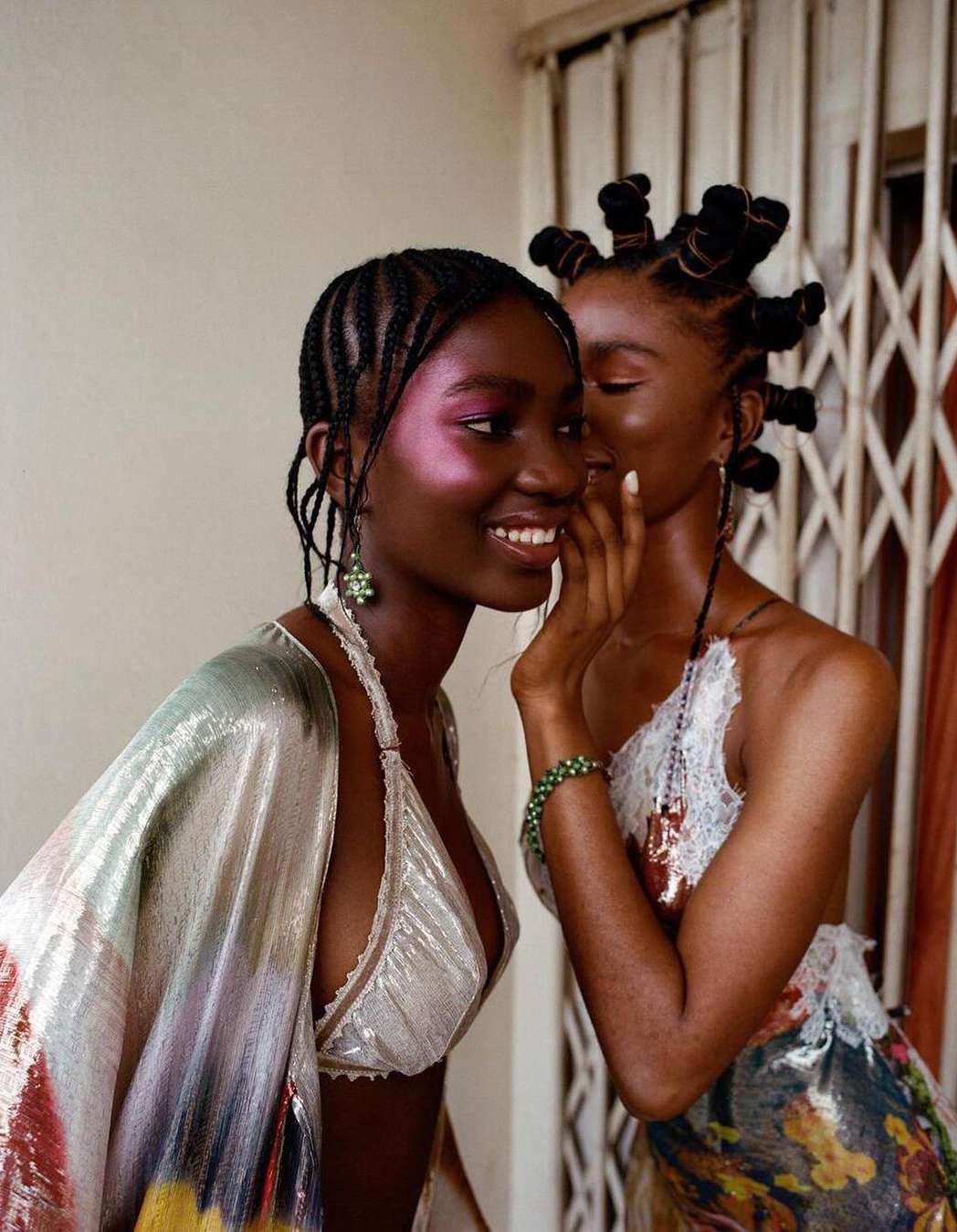 Two Black women in close portrait. One woman whispers into the other’s ear. They have elaborate braided hairstyles, and wear shiny tops.