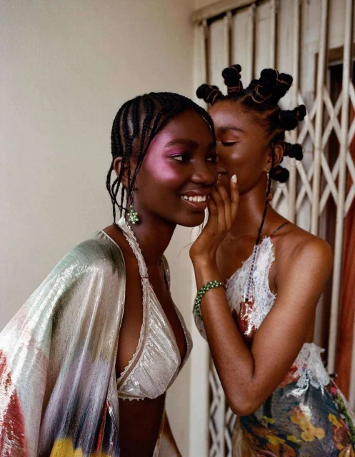 Two Black women in close portrait. One woman whispers into the other’s ear. They have elaborate braided hairstyles, and wear shiny tops.