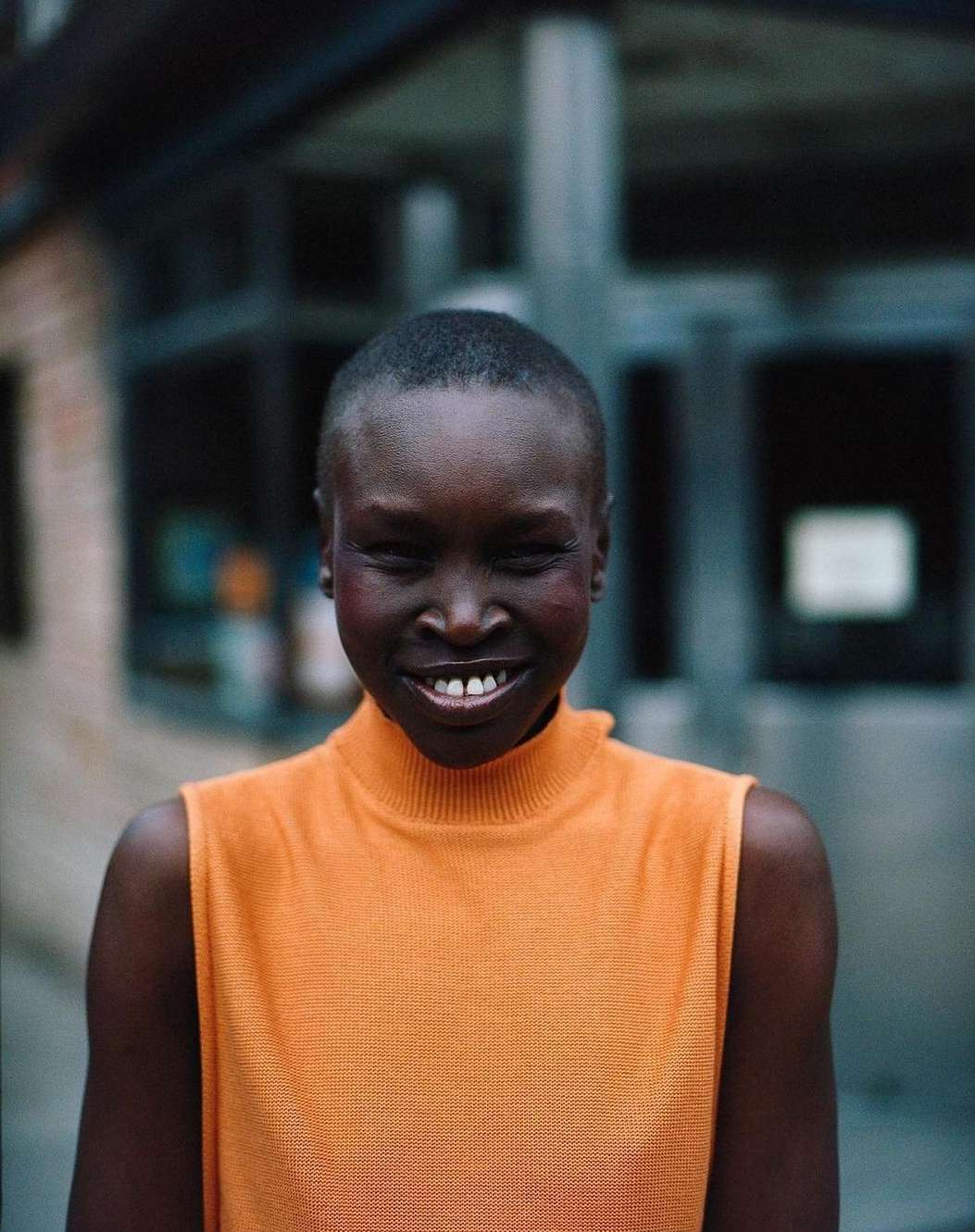 Portrait of a smiling adult woman with short hair wearing an orange top, with blurred urban background.