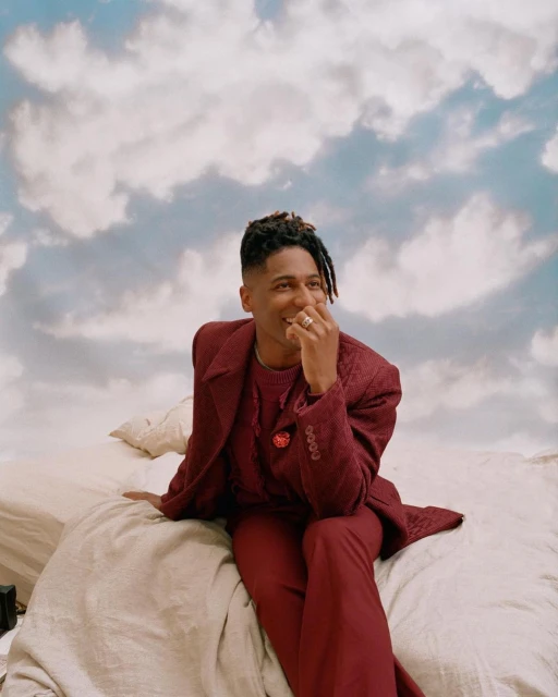 Stylish African American man in a burgundy suit sits on a bed in front of a cloudy sky backdrop, exuding a serene and contemplative mood.
