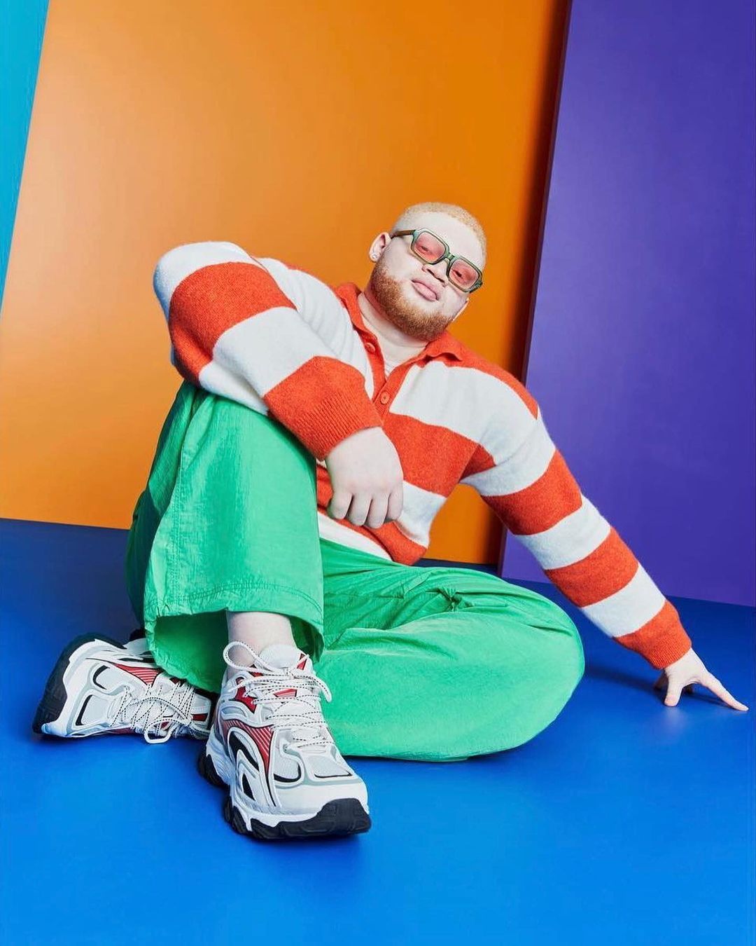Albino male model poses seated on a blue surface in the photo studio. He wears a striped shirt, green pants, and athletic shoes.