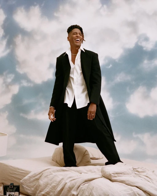 A smiling Black man in a white shirt and dark suit stands on a bed with a sky-themed backdrop, showcasing fashion and photographic artistry.