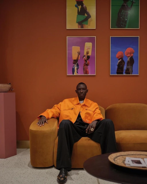 Adult man poses in an orange jacket and black pants, sitting in a tan chair against an orange wall decorated with framed artwork.