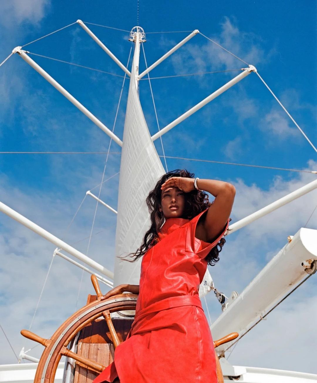 Woman in red dress stands at the helm of a white sailboat, shielding eyes, with blue sky and mast in background on a sunny day.