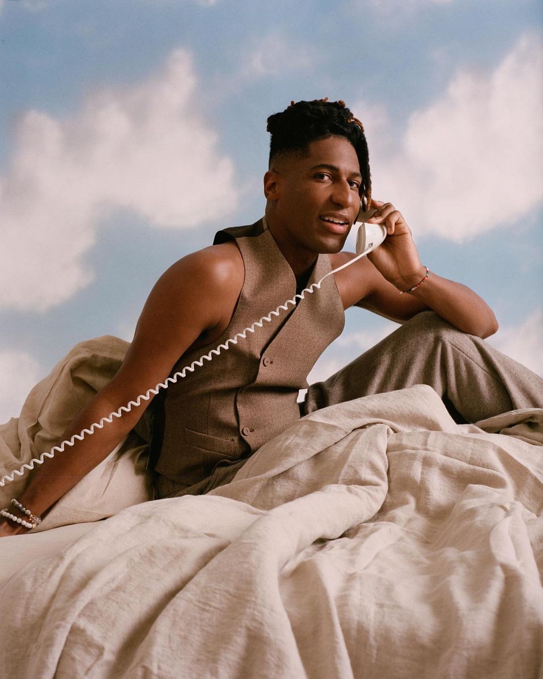 Portrait of a Black male in a vest and pants sitting in bed talking on the phone, set against a cloudy blue sky backdrop.