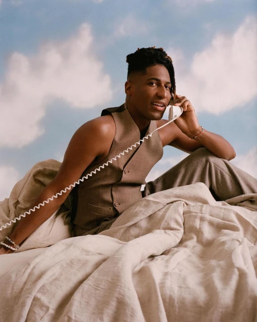 Portrait of a Black male in a vest and pants sitting in bed talking on the phone, set against a cloudy blue sky backdrop.