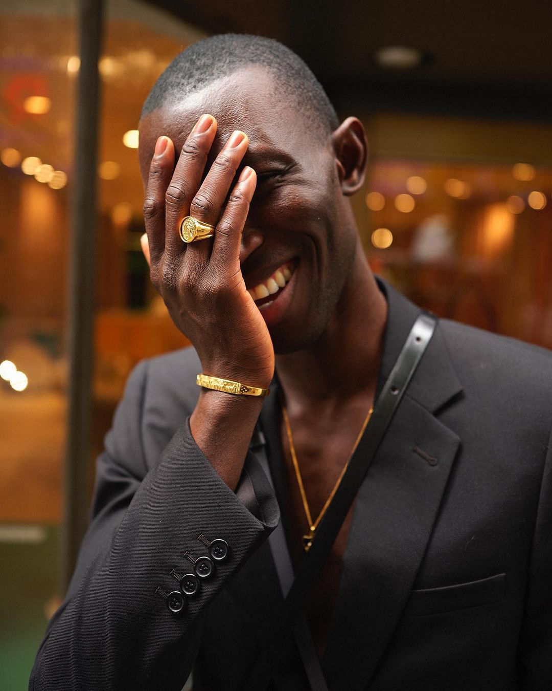 Smiling Black man wearing a black suit with gold jewellery including a bracelet and signet ring covering his face.