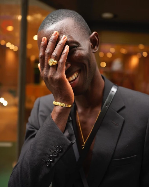 Smiling Black man wearing a black suit with gold jewellery including a bracelet and signet ring covering his face.