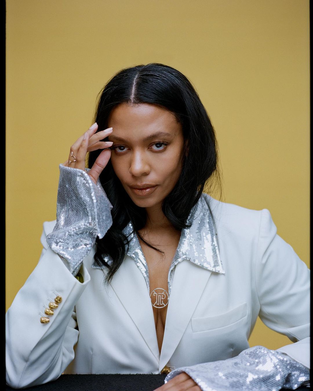 Headshot of Black female adult with hand near head wearing white blazer with metallic silver detailing against yellow backdrop in studio.