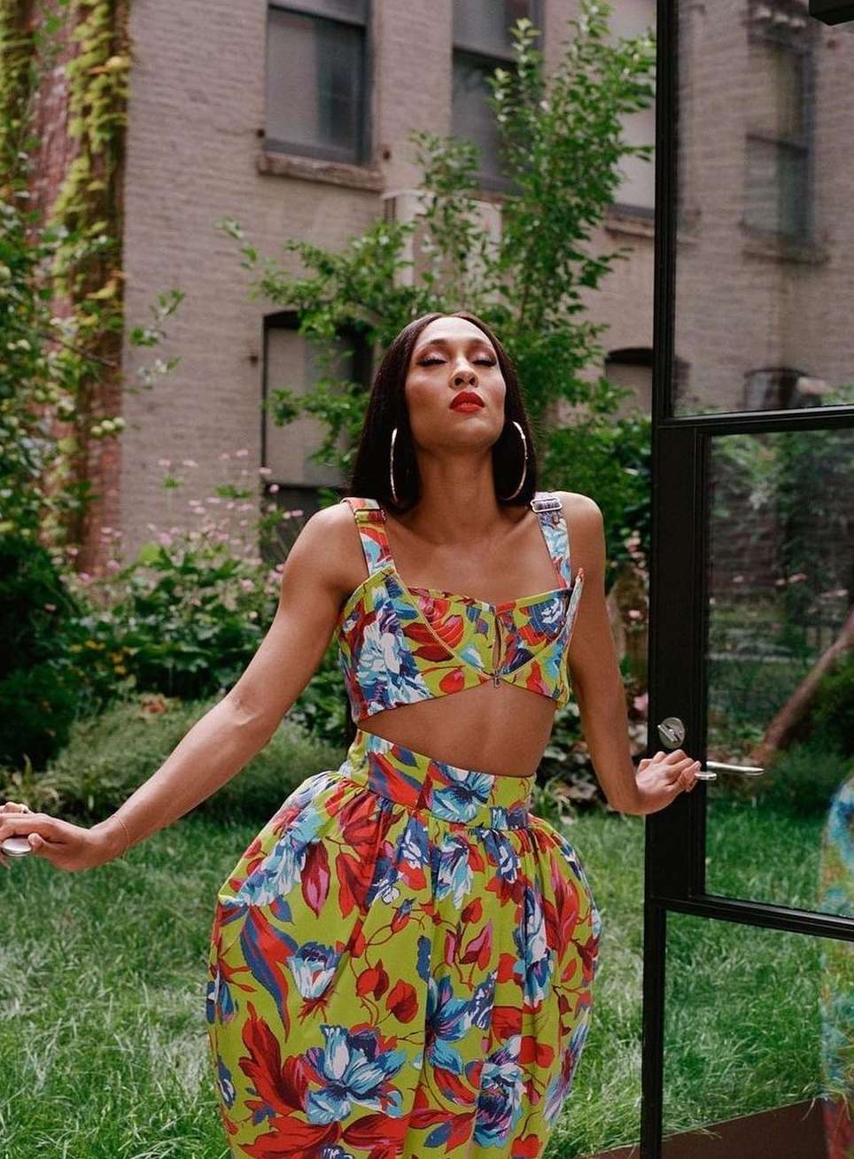 Elegant woman poses in a matching floral crop top and skirt in a lush garden setting with greenery and a brick building in the background.