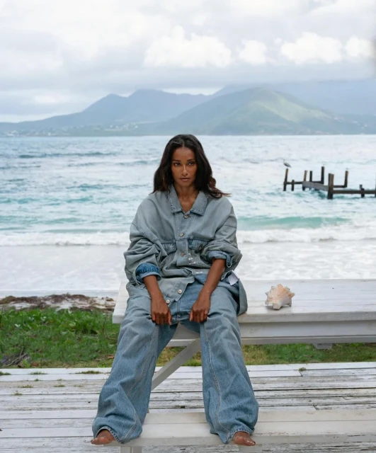 Woman in oversized denim sits on a white table on a beach with mountains in the background. A seashell sits on the table.