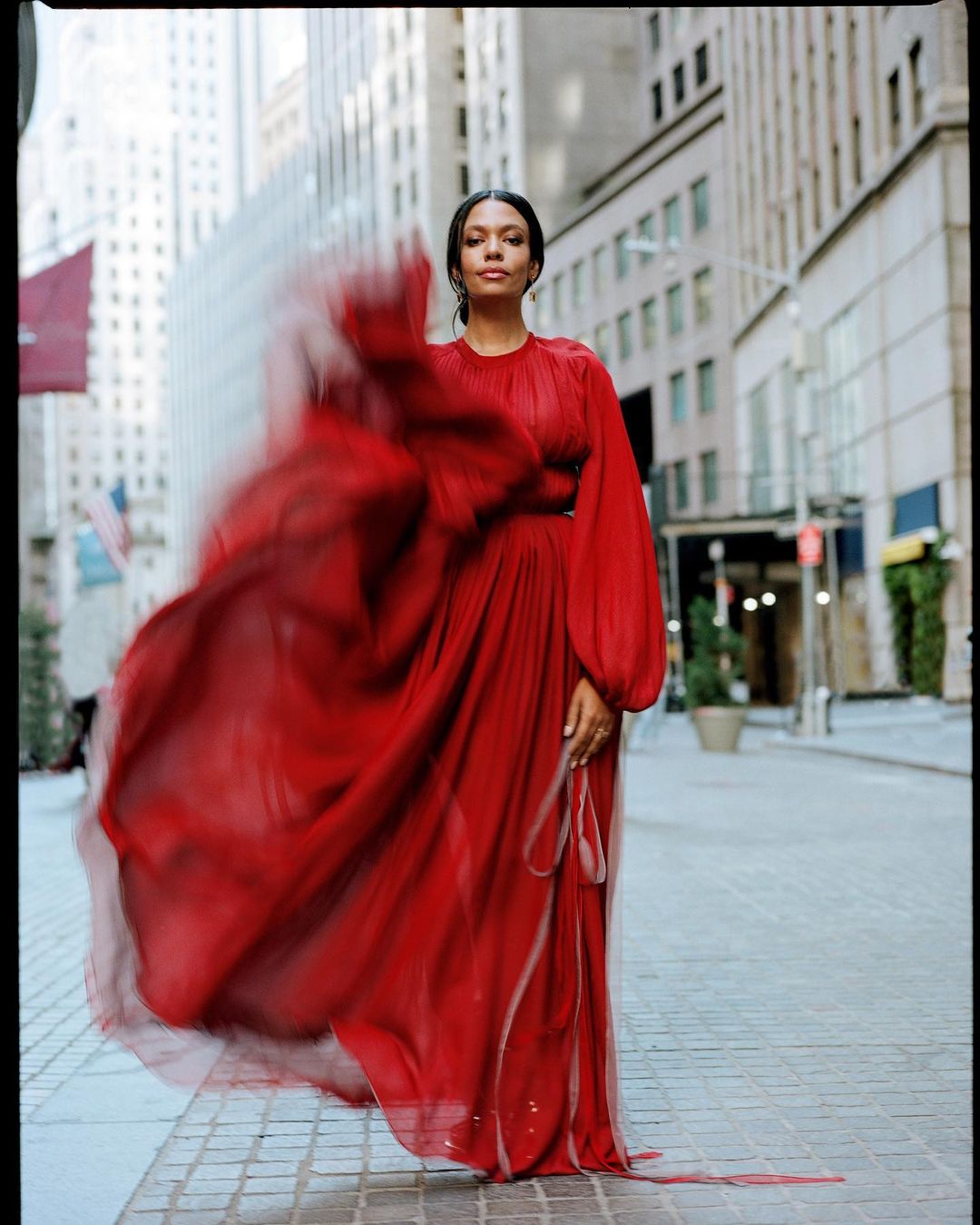 A woman in a red chiffon gown poses on a city street with tall buildings, creating a striking and vibrant portrait against an urban backdrop.