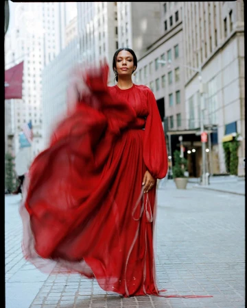A woman in a red chiffon gown poses on a city street with tall buildings, creating a striking and vibrant portrait against an urban backdrop.