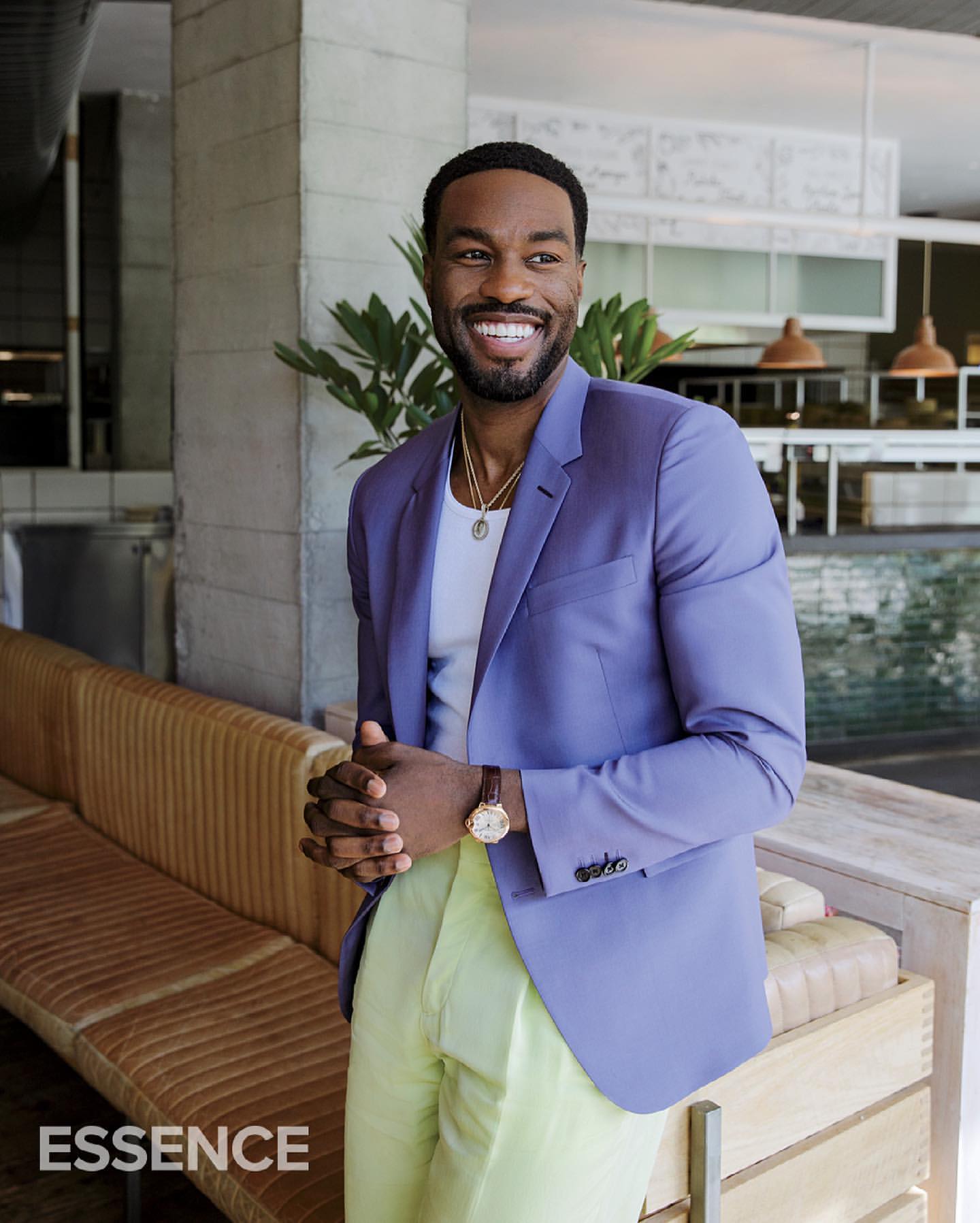 Smiling Black man in a light purple blazer and yellow pants posing indoors next to a couch in front of a restaurant.