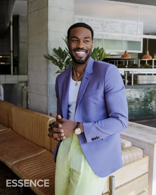 Smiling Black man in a light purple blazer and yellow pants posing indoors next to a couch in front of a restaurant.
