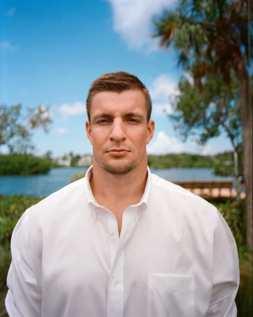 Portrait of a white man in a white button-down shirt outdoors against a body of water and palm trees under a bright blue sky.