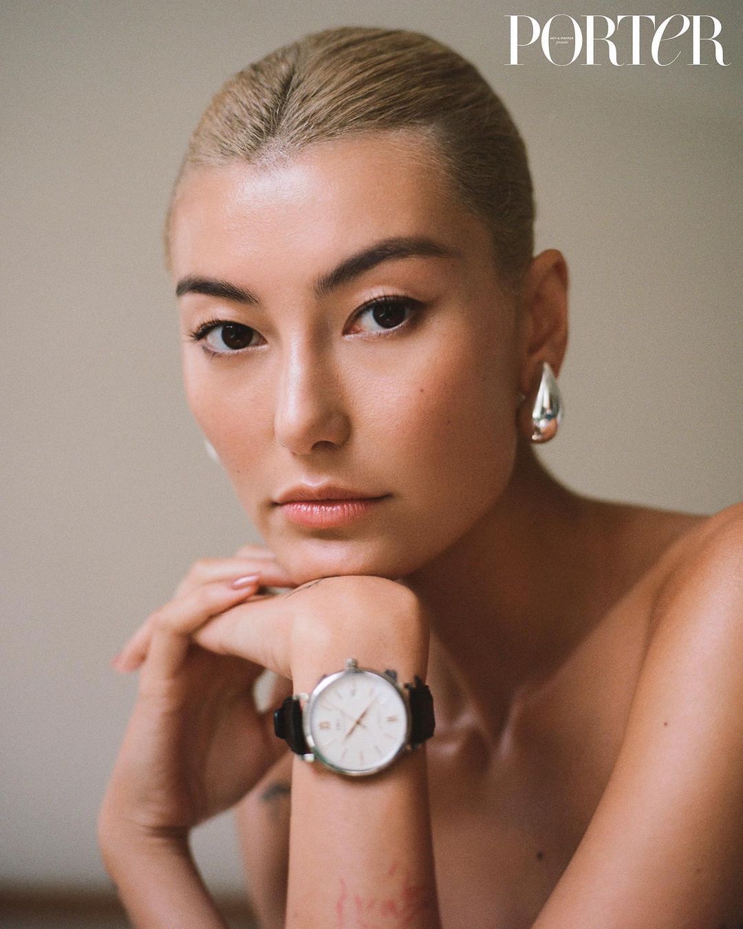 Portrait of a woman with blonde hair wearing a wristwatch and silver drop earrings, indoors with neutral wall and natural lighting.