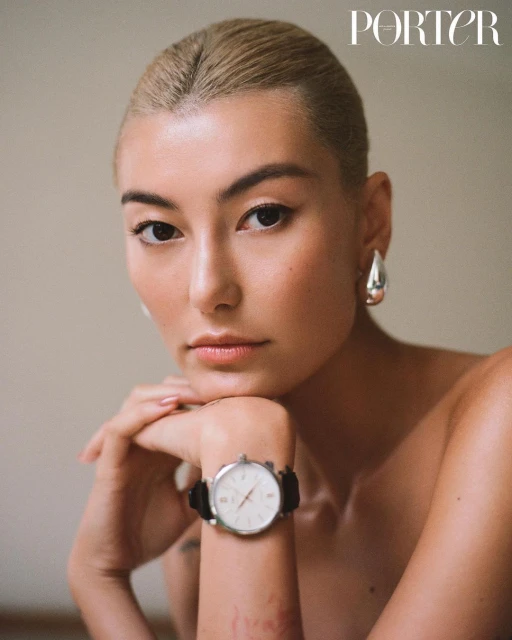 Portrait of a woman with blonde hair wearing a wristwatch and silver drop earrings, indoors with neutral wall and natural lighting.