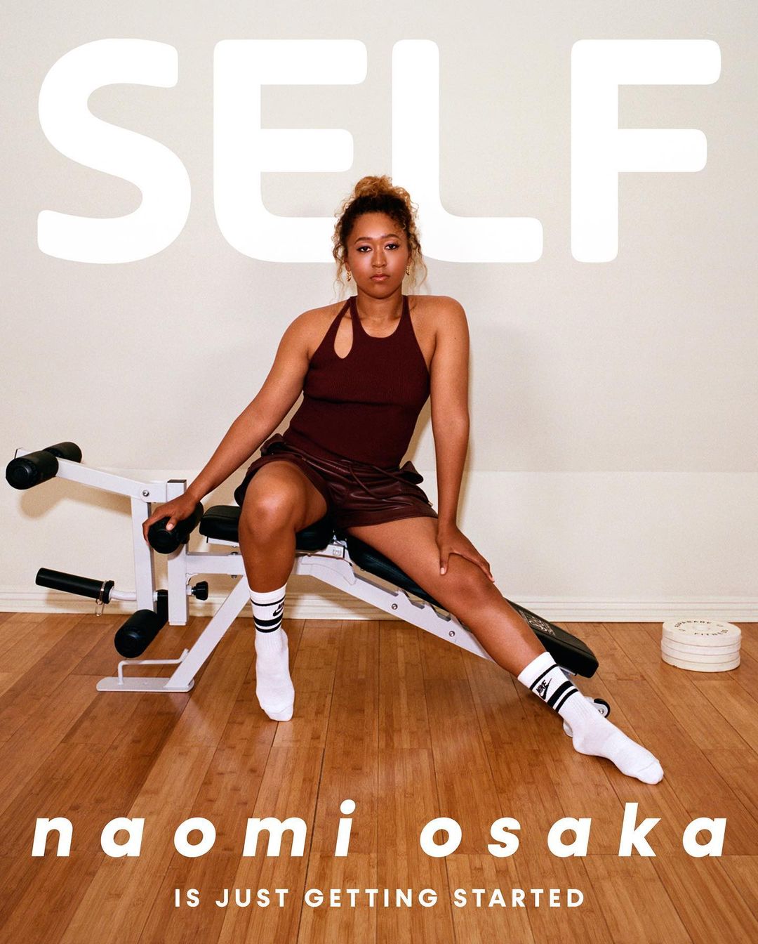 Naomi Osaka sits on a workout bench in a fitness photoshoot for Self Magazine. The workout is in a light-filled indoor setting.