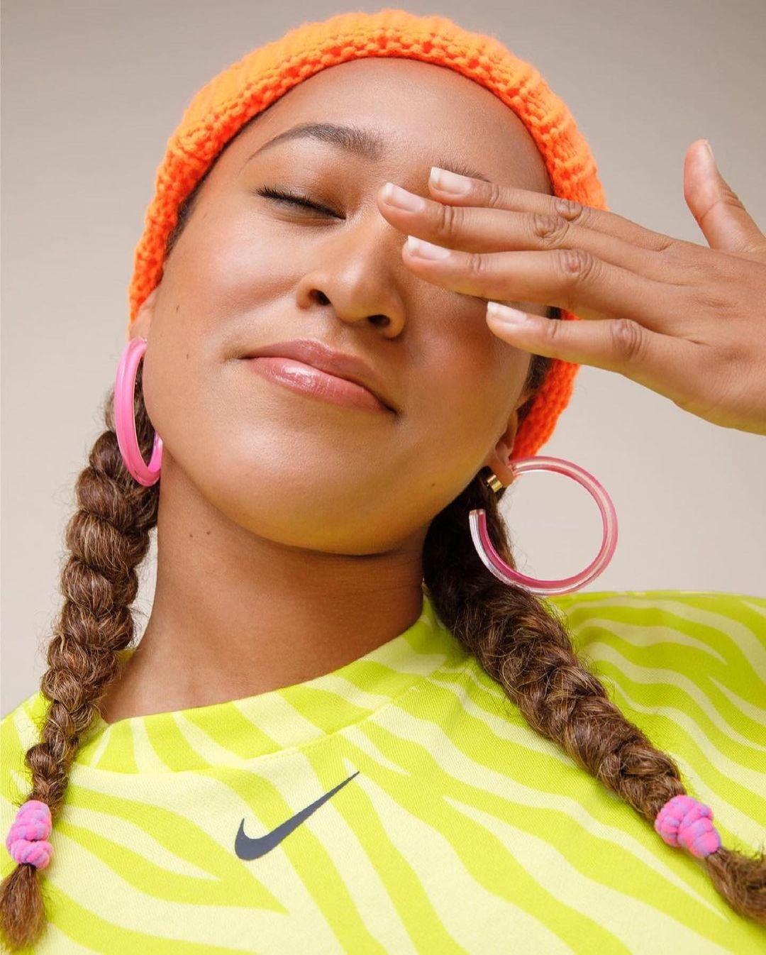 Close-up portrait of an adult woman with braided hair wearing a bright orange knit hat and pink hoop earrings, covering one eye with hand.