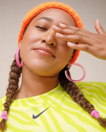 Close-up portrait of an adult woman with braided hair wearing a bright orange knit hat and pink hoop earrings, covering one eye with hand.