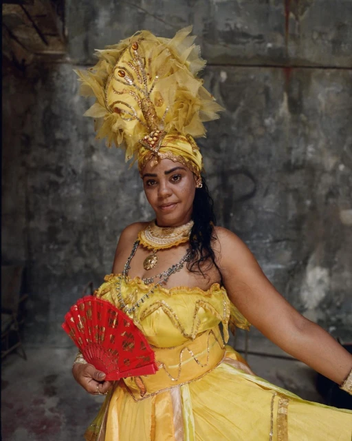 Portrait of an adult woman wearing a yellow costume with a feathered headdress, holding a red fan, against a grey wall backdrop.