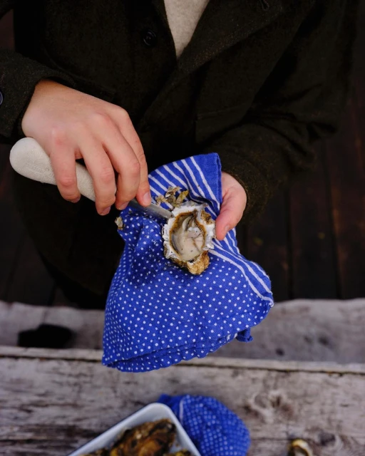 Person shucking an oyster with oyster knife on blue polka dot towel. Rustic wooden table and oysters in white dish.