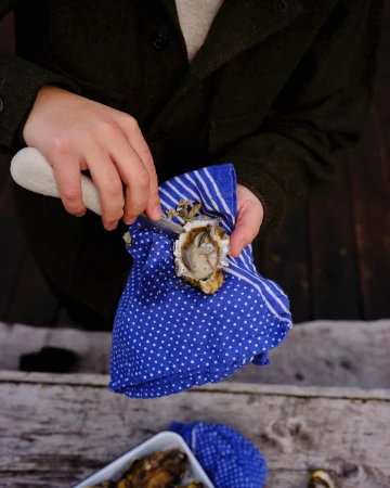 Person shucking an oyster with oyster knife on blue polka dot towel. Rustic wooden table and oysters in white dish.