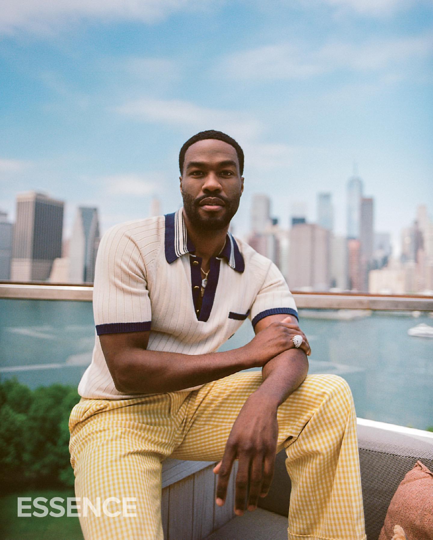 Portrait of a Black man in a collared shirt and trousers, seated outdoors against an urban skyline on a sunny day.