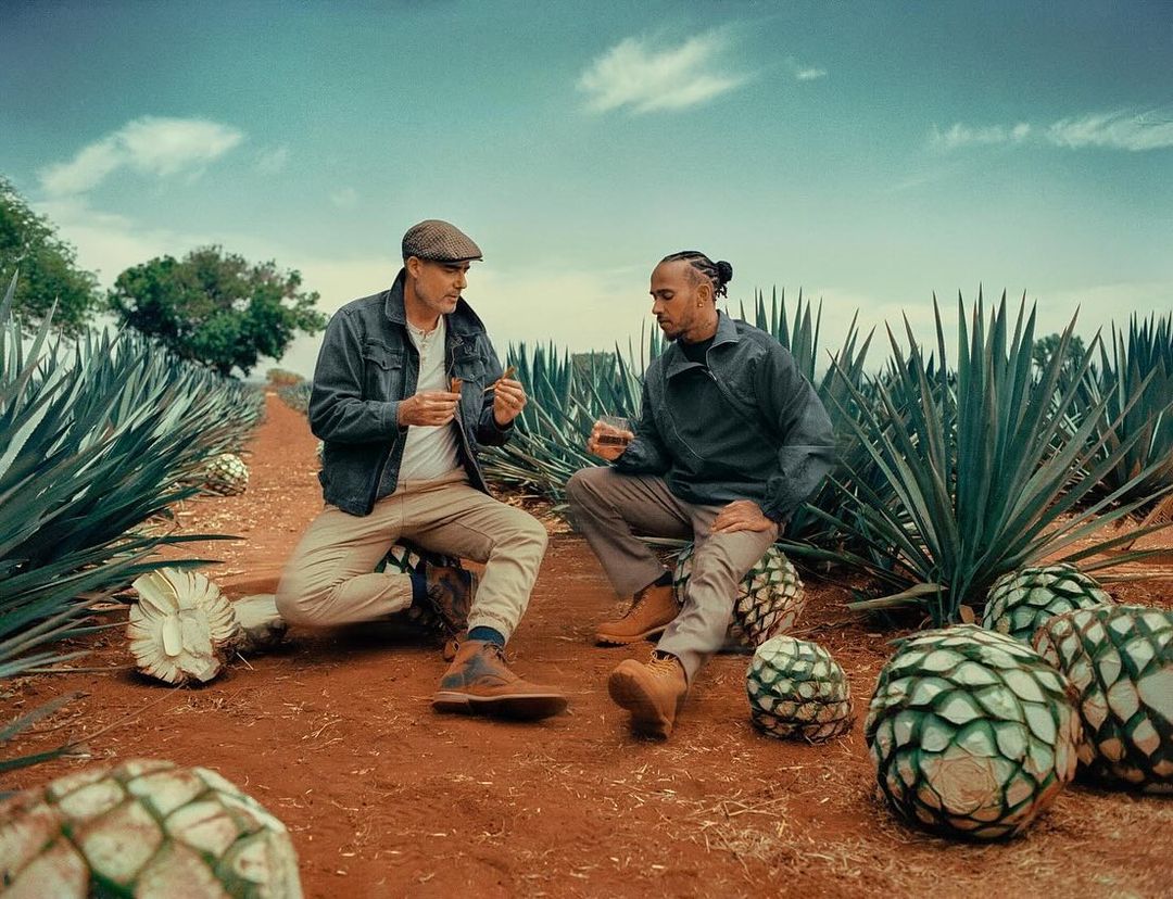 Two men sit among agave plants in a field, one holding a glass, the other holding cigars, in a setting suggestive of a beverage brand.