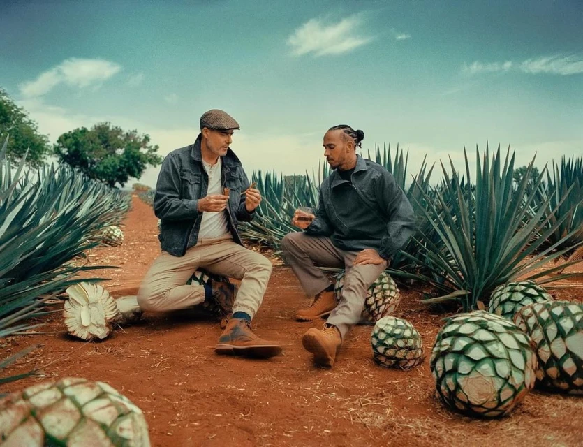 Two men sit among agave plants in a field, one holding a glass, the other holding cigars, in a setting suggestive of a beverage brand.