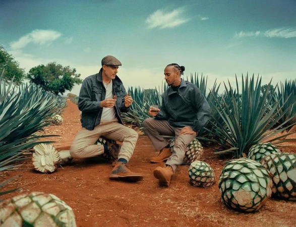 Two men sit among agave plants in a field, one holding a glass, the other holding cigars, in a setting suggestive of a beverage brand.