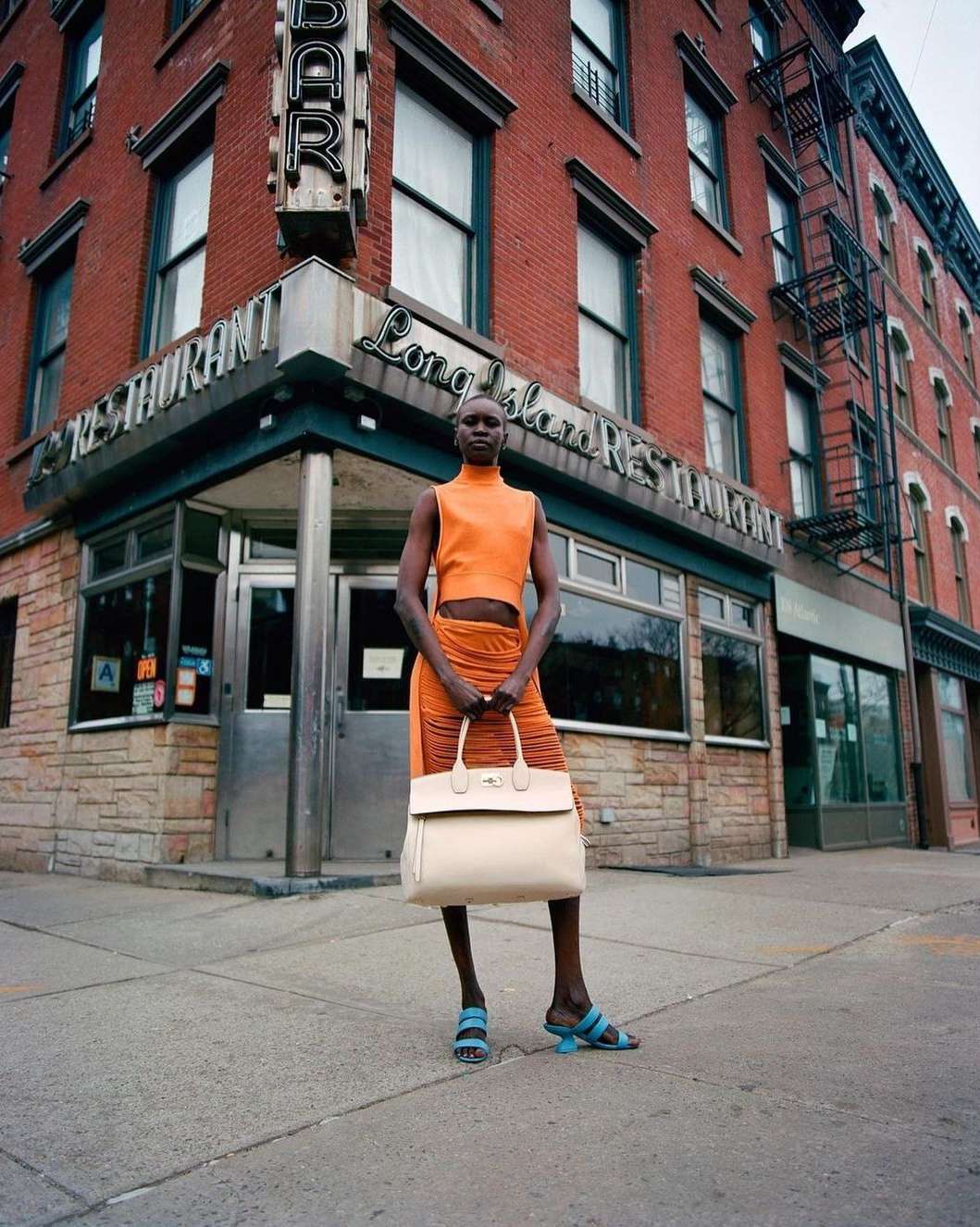 Woman in orange outfit stands in front of brick building with 'Long Island Restaurant' sign, holding a handbag on a city street.