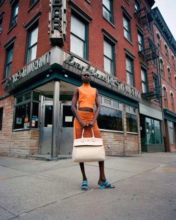 Woman in orange outfit stands in front of brick building with 'Long Island Restaurant' sign, holding a handbag on a city street.