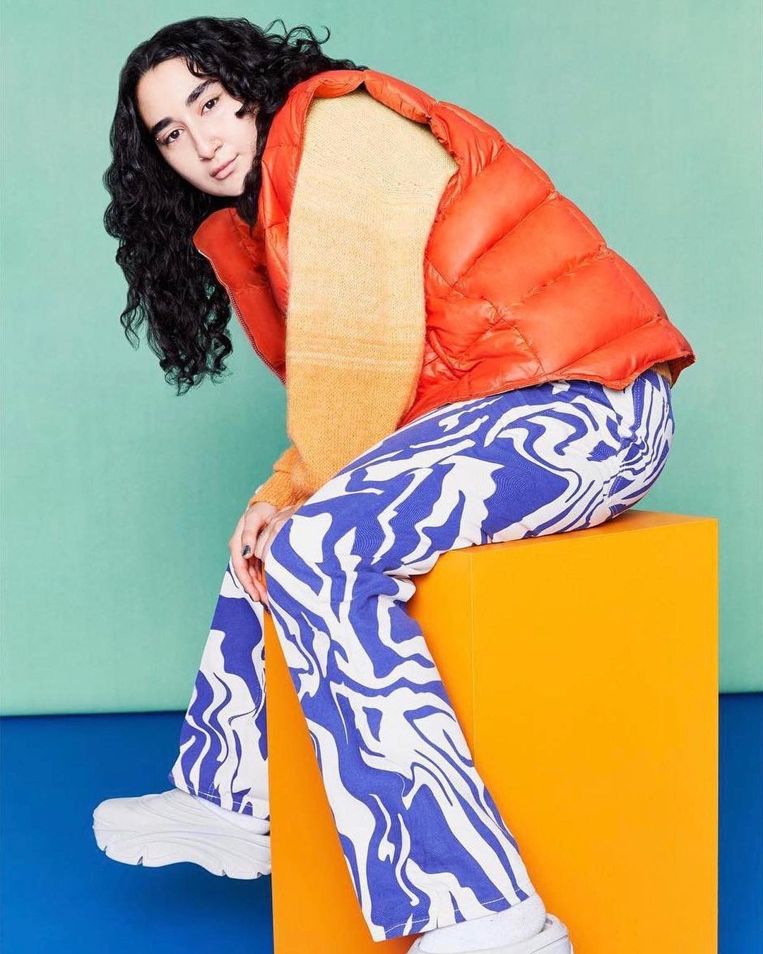 A person sits on an orange cube in a studio setting, wearing a puffer vest, patterned pants, and white sneakers in a colourful setting.