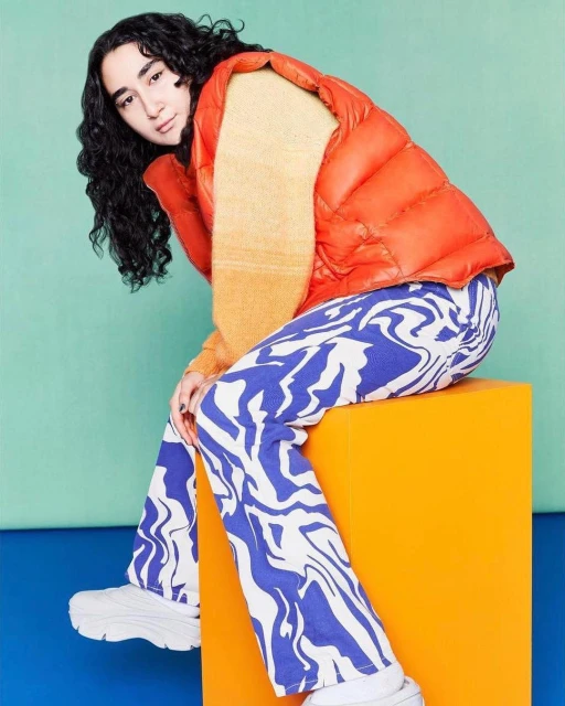 A person sits on an orange cube in a studio setting, wearing a puffer vest, patterned pants, and white sneakers in a colourful setting.