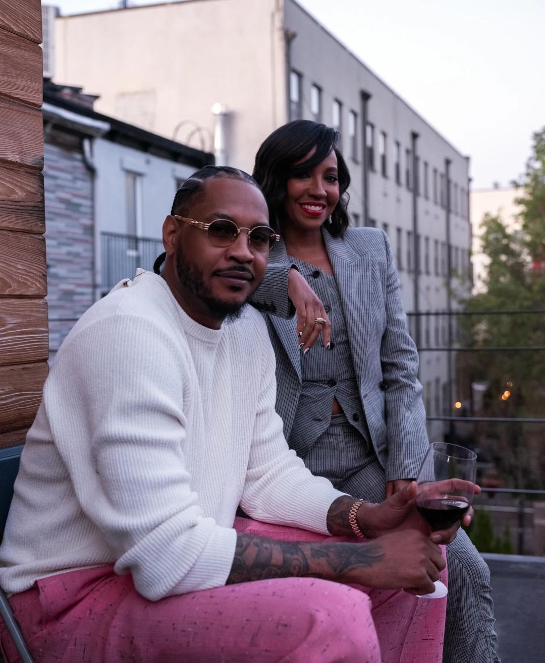 Fashionable couple poses outdoors in urban setting. Man in pink pants holds wine; woman in grey suit leans on his shoulder.