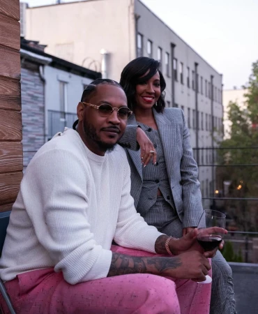 Fashionable couple poses outdoors in urban setting. Man in pink pants holds wine; woman in grey suit leans on his shoulder.