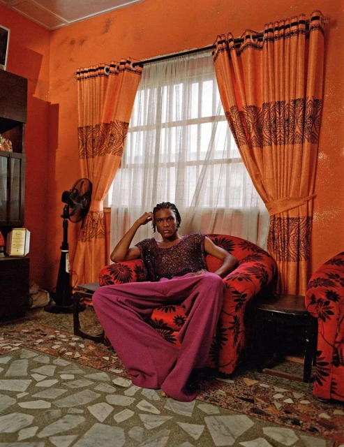 Person in purple pants sits in a red armchair in an orange room. There is a standing fan, patterned curtains, and a patterned floor.