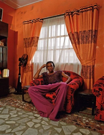Person in purple pants sits in a red armchair in an orange room. There is a standing fan, patterned curtains, and a patterned floor.