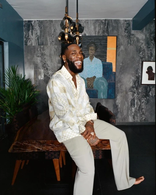Burna Boy is pictured indoors, seated on a wooden table with a wide smile; wearing a patterned shirt and light pants with a painting in background.