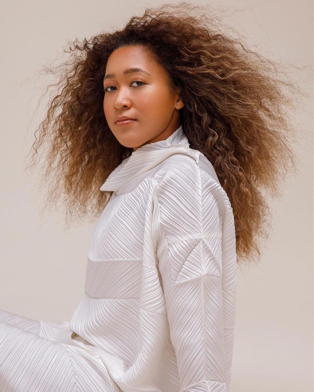 Portrait of Naomi Osaka in a white, pleated, long-sleeve shirt, with a neutral background, showcasing her curly hair and serene expression.