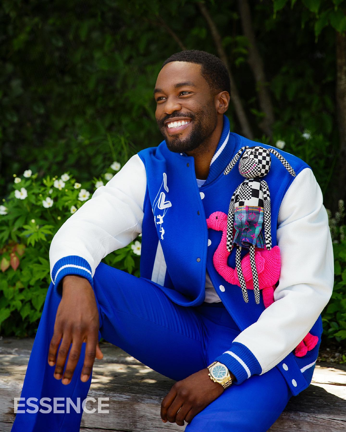 Smiling Black man in blue suit jacket and pants poses outdoors with a plush flamingo and rabbit decoration pinned to his jacket.